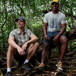 Two men in casual outdoor wear and caps smile on tree branches in a lush forest, their Free Country Men's Super Soft Microtech® Short Sleeve Graphic Crew Tees keeping them cool and comfortable amid dense foliage.