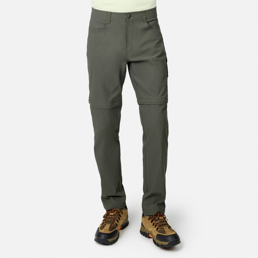 Lower body visible: person wears Free Country Men’s Nylon Stretch Convertible Pant in olive green—four-way stretch, water-repellent, converts to shorts via below-knee zippers—paired with brown hiking boots. Plain white background.