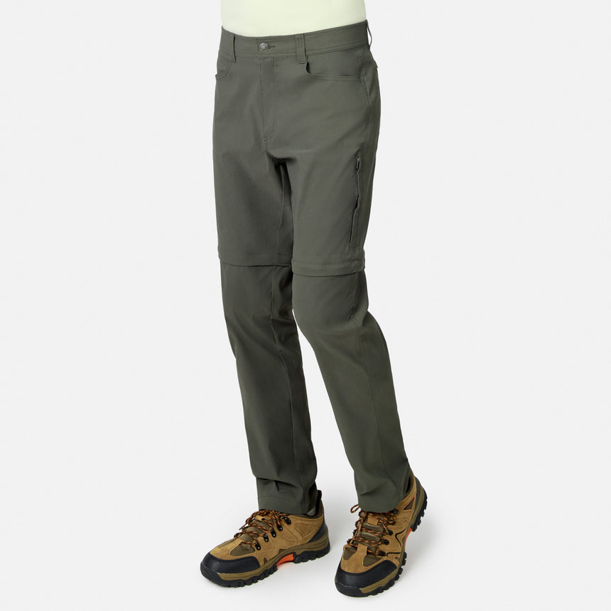 A man wearing Free Country Men’s Nylon Stretch Convertible Pants in olive-green with zip-off legs and brown hiking boots, shown from the waist down against a plain white background.