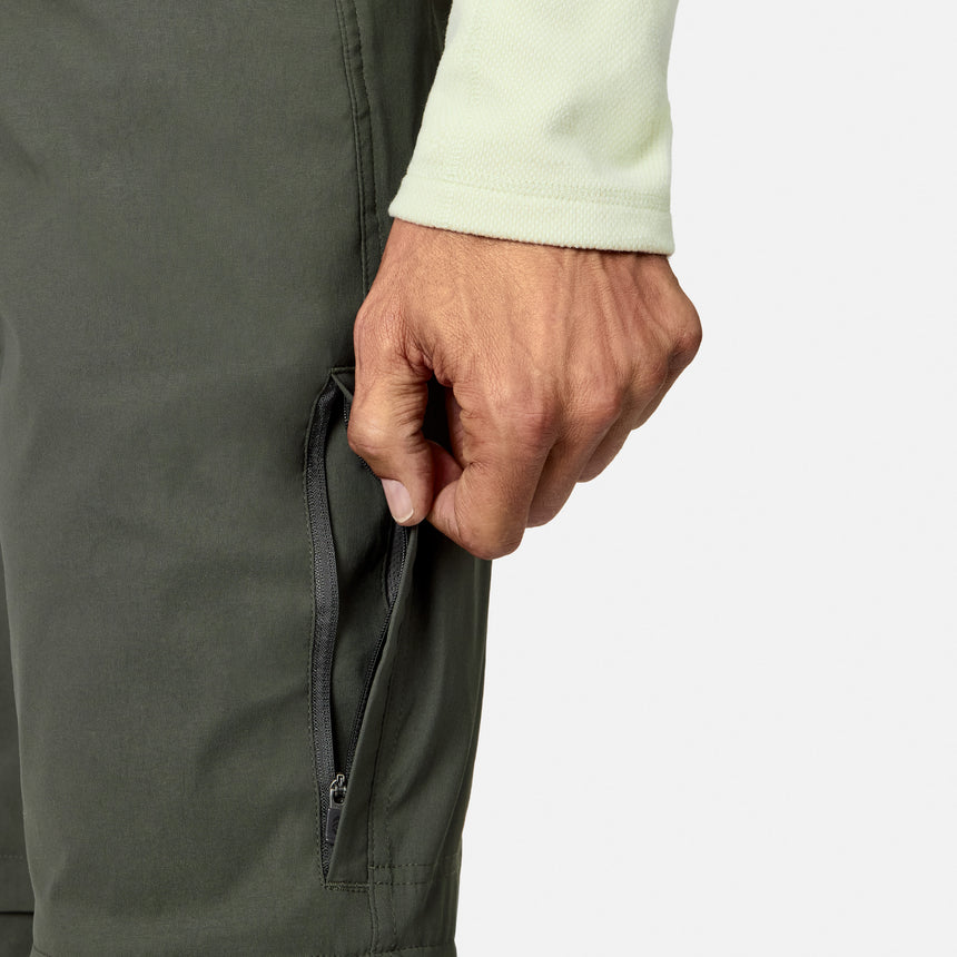 A person wearing a light long-sleeve shirt opens a zippered pocket on Free Country Men’s Nylon Stretch Convertible Pant in dark green, with just their hand, part of the arm, and the pocket visible against a plain background.