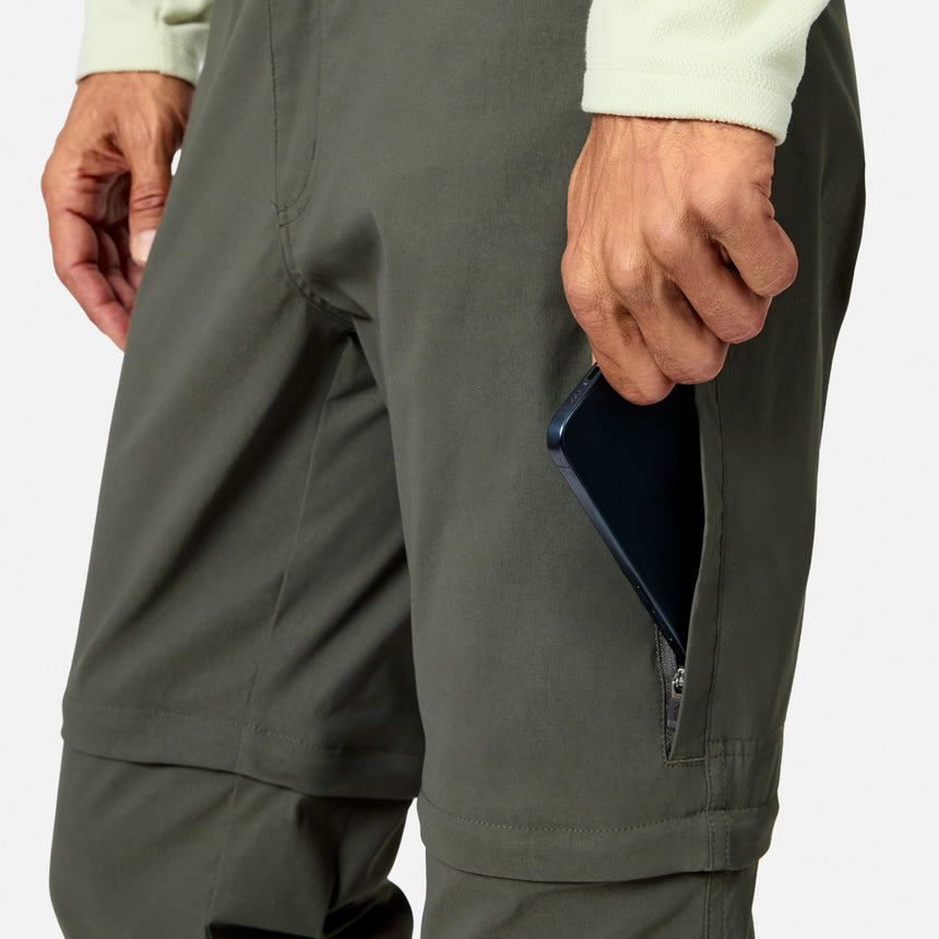 A man in Free Country Men’s Nylon Stretch Convertible Pant in olive stows a black smartphone into the zippered side pocket; only his hand and lower torso are shown.