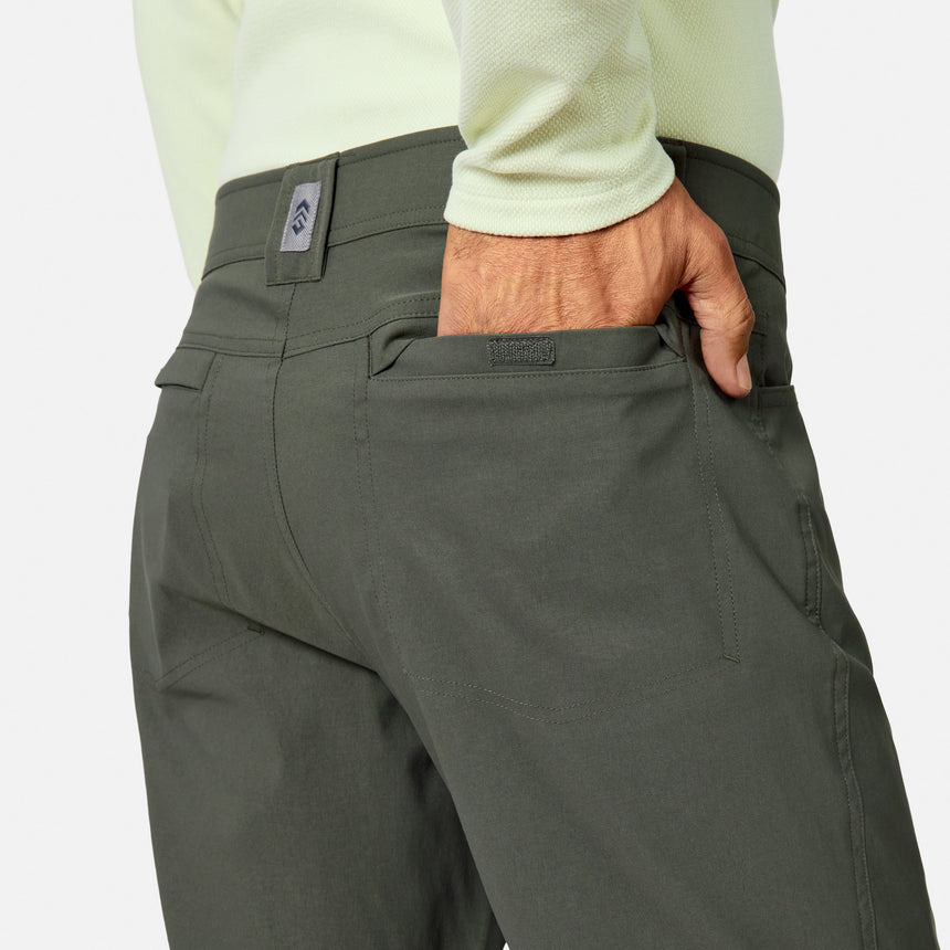A person in Free Country Men’s Nylon Stretch Convertible Pants (dark green, black logo above back pocket) and a light green long-sleeve shirt puts their right hand in the back pocket.