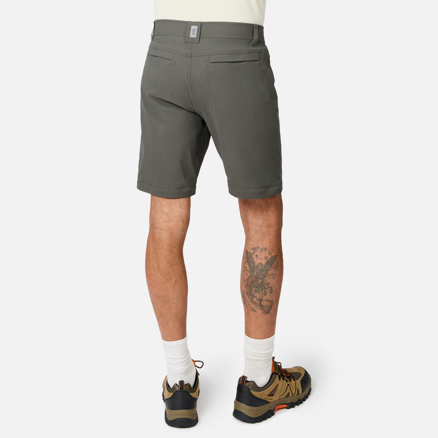 A person faces away in Free Country Men’s Nylon Stretch Convertible Pant, white socks, brown hiking shoes, and a light shirt. A tattoo is visible on the left calf. The background is plain and light-colored.