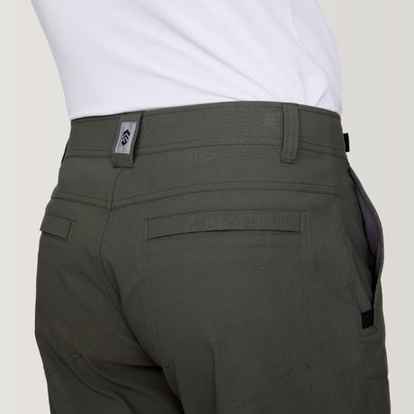 [Model is wearing a size Medium.] Man wearing Free Country Men's Nylon Stretch Trek Short in Olive, size M, made from lightweight stretch performance fabric featuring a water repellent finish, an adjustable waist belt, two back welt pockets, and two side sipper pockets, styled with a white t shirt and white shoes #color_olive