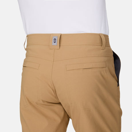 A person is shown from the back wearing Free Country Men's Nylon Stretch Trek Shorts in tan and a white shirt, highlighting the shorts' adjustable waist belt, waistband, belt loops, and rear pockets.