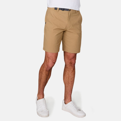 A person is seen from the waist down, wearing Free Country Men's Nylon Stretch Trek Shorts in tan with an adjustable waist belt and white sneakers, standing against a plain white background. The upper body is not visible.