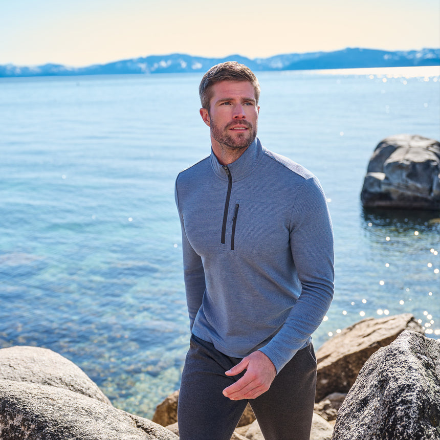 A man wearing the Free Country Men’s Altitude Quilt 1/2 Zip Mock Neck Shirt in light blue stands on a rocky shore beside a clear lake, with snow-capped mountains and a sunny sky in the distance.