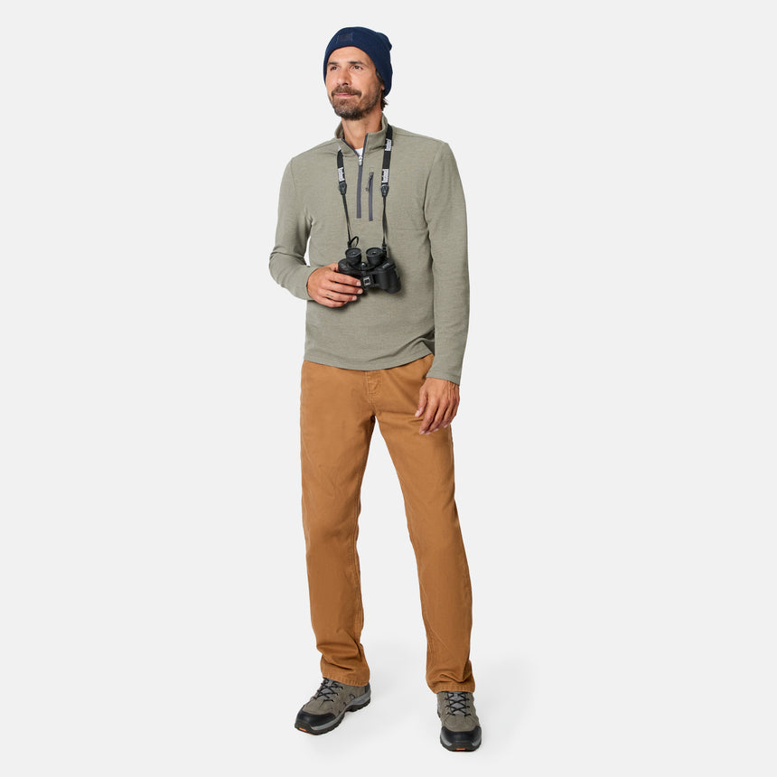 A man wearing a blue beanie, brown pants, hiking shoes, and the Free Country Men’s Altitude Quilt 1/2 Zip Mock Neck Shirt in gray stands holding binoculars and looking slightly to his left.