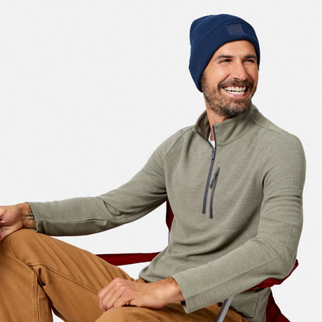 A man with a beard smiles while sitting in a chair, wearing the Free Country Men’s Altitude Quilt 1/2 Zip Mock Neck Shirt in olive green, along with a blue beanie and tan pants. The background is plain and light-colored.