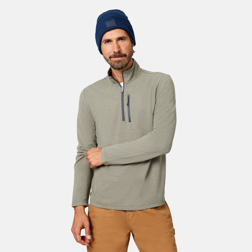 A man with a beard wears a blue beanie, tan pants, and the Free Country Men’s Altitude Quilt 1/2 Zip Mock Neck Shirt in Forest Heather, which features comfortable 4-way stretch for easy movement. He stands against a plain light background.