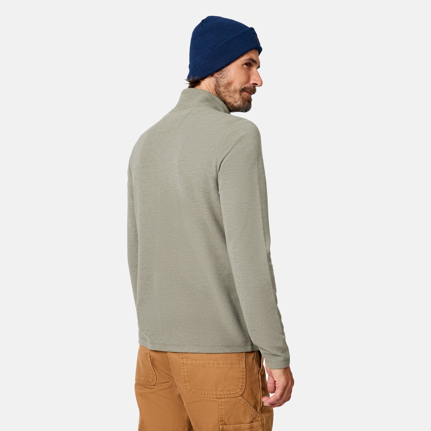 A man wearing the Free Country Men’s Altitude Quilt 1/2 Zip Mock Neck Shirt in light gray, paired with a blue beanie and brown pants, stands with his back turned and looks over his shoulder against a plain white background.