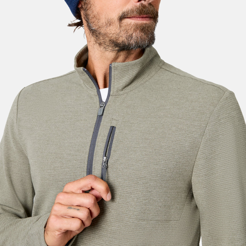 A man, shown from the lower face to upper torso, wears the Free Country Men’s Altitude Quilt 1/2 Zip Mock Neck Shirt—a green lightweight quilted top with a gray half-zip and chest pocket—paired with a navy beanie and is partially unzipping the shirt.