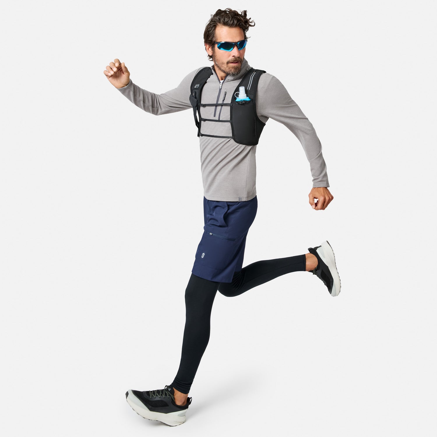 A man wearing sunglasses, the Free Country Men’s Altitude Quilt 1/2 Zip Mock Neck Shirt in gray, navy shorts, black leggings, and running shoes is running. He has on a black hydration vest and carries a collapsible blue water bottle in a chest pocket.