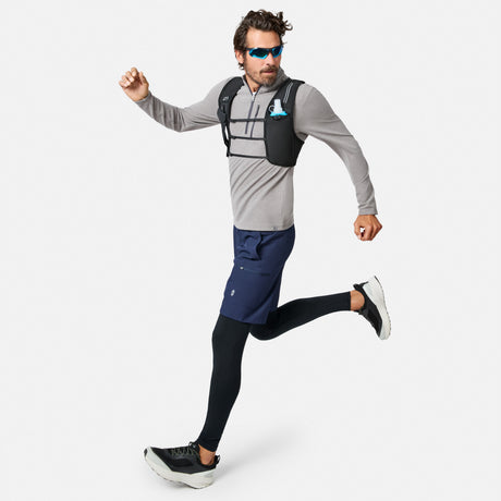 A man wearing sunglasses, the Free Country Men’s Altitude Quilt 1/2 Zip Mock Neck Shirt in gray, navy shorts, black leggings, and running shoes is running. He has on a black hydration vest and carries a collapsible blue water bottle in a chest pocket.