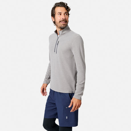 A man with wavy brown hair and a beard stands smiling, looking to his right against a plain white background, wearing the Free Country Men’s Altitude Quilt 1/2 Zip Mock Neck Shirt in light gray, paired with navy blue shorts and black leggings.