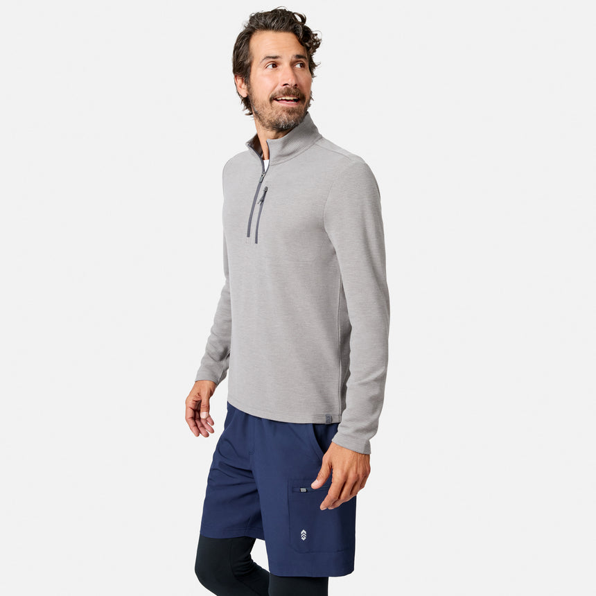 A man with wavy brown hair and a beard stands smiling, looking to his right against a plain white background, wearing the Free Country Men’s Altitude Quilt 1/2 Zip Mock Neck Shirt in light gray, paired with navy blue shorts and black leggings.