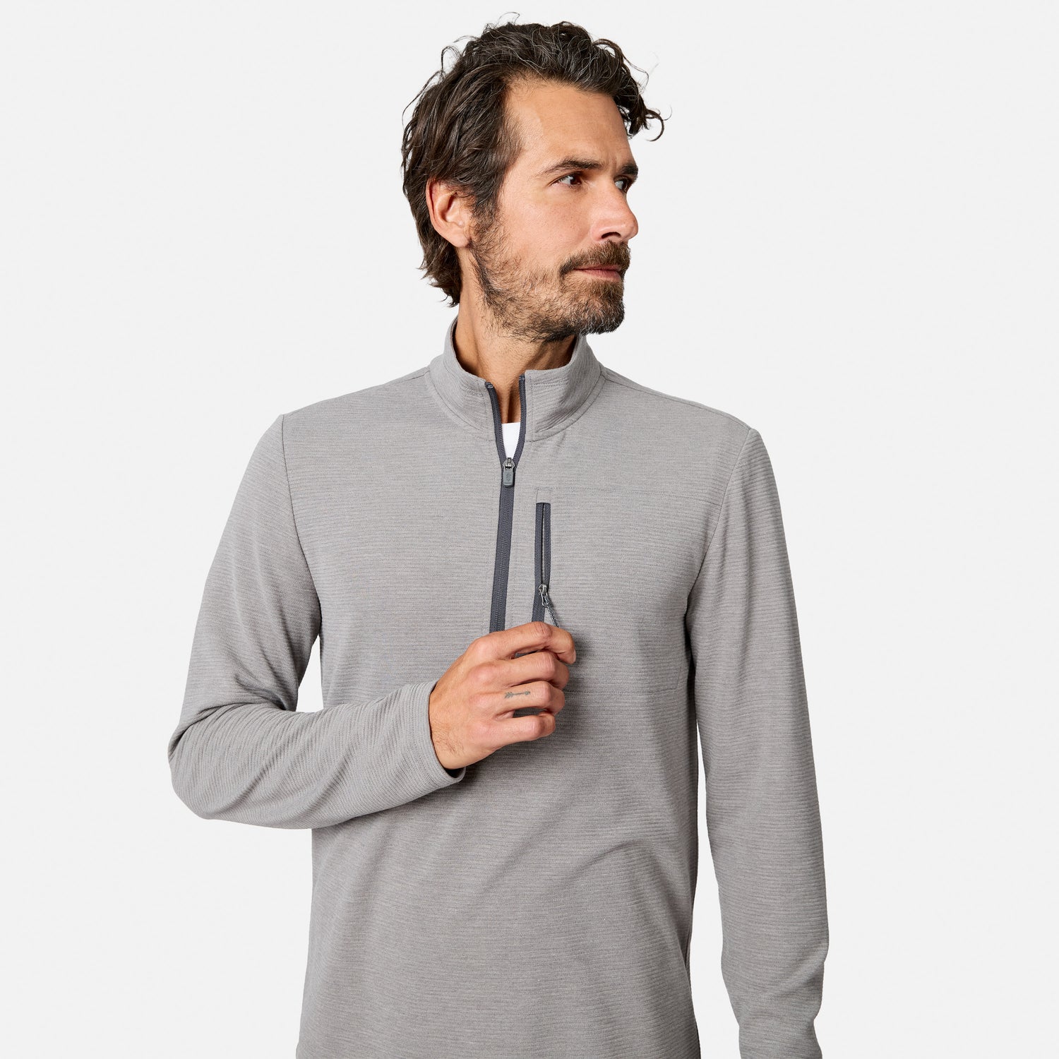 A man with dark hair and a beard wears the Free Country Men’s Altitude Quilt 1/2 Zip Mock Neck Shirt in Grey Heather. He looks to his left while holding the zipper with his right hand against a plain white background.