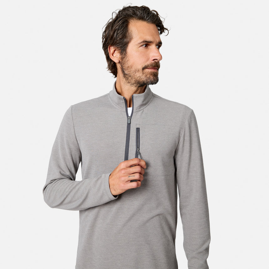 A man with dark hair and a beard wears the Free Country Men’s Altitude Quilt 1/2 Zip Mock Neck Shirt in Grey Heather. He looks to his left while holding the zipper with his right hand against a plain white background.