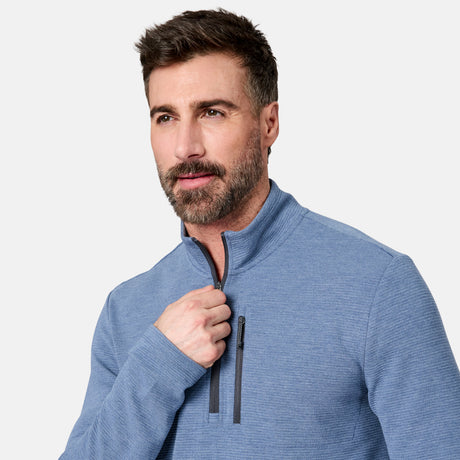 A man with short brown hair and a beard wears the Free Country Men’s Altitude Quilt 1/2 Zip Mock Neck Shirt in light blue. Looking slightly to the side, he is pulling up the zipper of the pullover, which features 4-way stretch for comfort. The shirt stands out against a plain light gray background.
