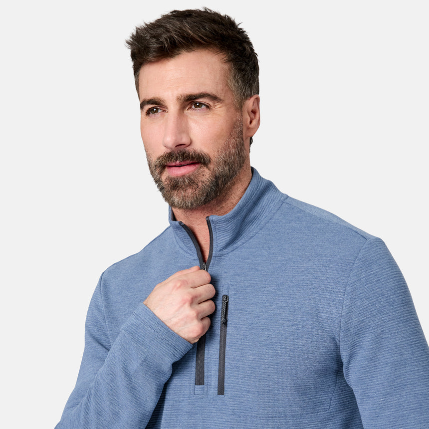 A man with short brown hair and a beard wears the Free Country Men’s Altitude Quilt 1/2 Zip Mock Neck Shirt in light blue. Looking slightly to the side, he is pulling up the zipper of the pullover, which features 4-way stretch for comfort. The shirt stands out against a plain light gray background.
