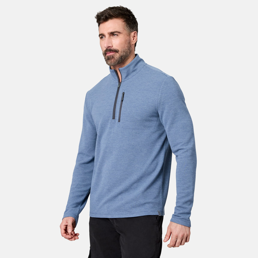 A man with short brown hair and a beard stands against a plain white background, wearing the Free Country Men’s Altitude Quilt 1/2 Zip Mock Neck Shirt in light blue, which features a zippered chest pocket, along with black pants.