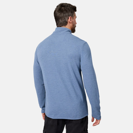 A man with short brown hair is standing with his back to the camera, wearing the Free Country Men’s Altitude Quilt 1/2 Zip Mock Neck Shirt in light blue and black pants against a plain white background.