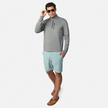 A man in sunglasses and a gray pullover stands confidently in Clearance Men’s Trek Short—light blue, four-way stretch, lightweight, and breathable—against a plain white background.
