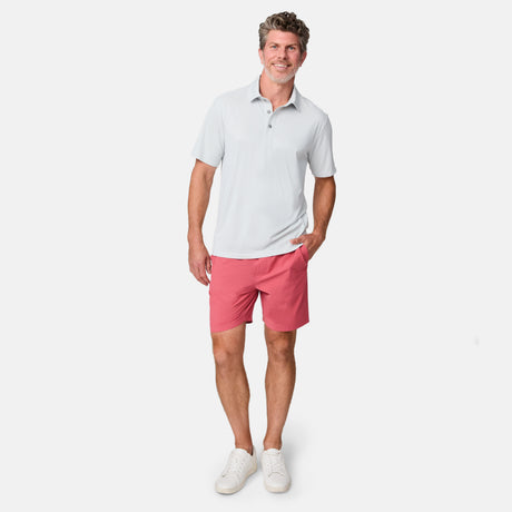 Smiling man wearing a light gray polo, Clearance Men’s Trek Short in pink, and white sneakers stands with one hand in his pocket against a plain white background.