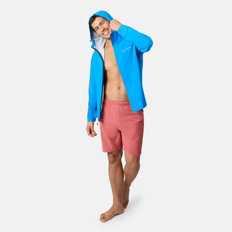 A smiling man stands barefoot against a plain light background, wearing Clearance Men’s Trek Short and a bright blue hooded jacket.