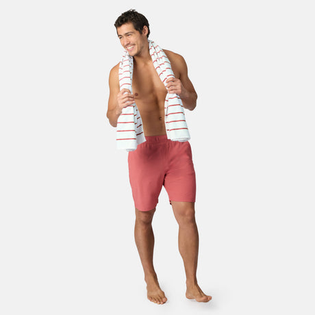 A smiling man stands barefoot in Clearance Men’s Trek Short swim trunks—lightweight, breathable red shorts—and a white towel with red stripes draped around his neck against a plain white background.