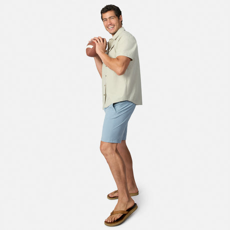A man in a short-sleeve button-up shirt, Clearance Men’s Trek Short, and sandals holds a football in a throwing stance, smiling against a plain white background.