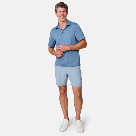 A man smiles in a blue polo, Clearance Men’s Trek Short, and white sneakers, holding a white cup against a plain background. His Trek Shorts feature breathable fabric for all-day comfort.