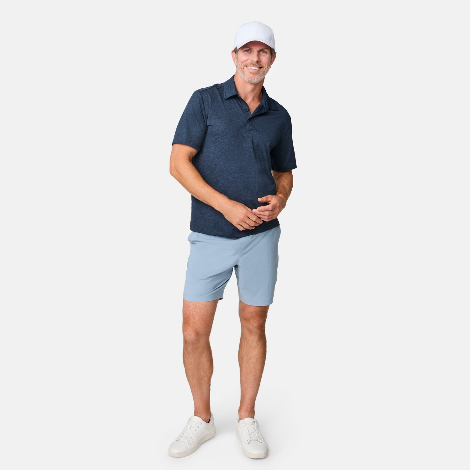 A man in a white cap, navy polo, Clearance Men’s Trek Short, and white sneakers stands smiling against a plain white background.