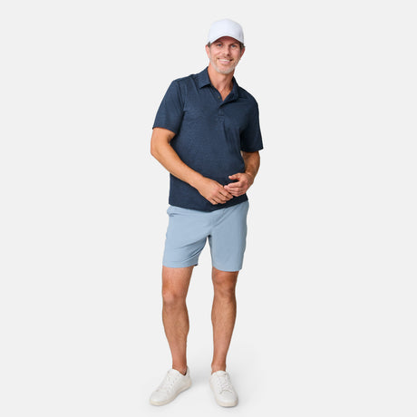 A man in a white cap, navy polo, Clearance Men’s Trek Short, and white sneakers stands smiling against a plain white background.