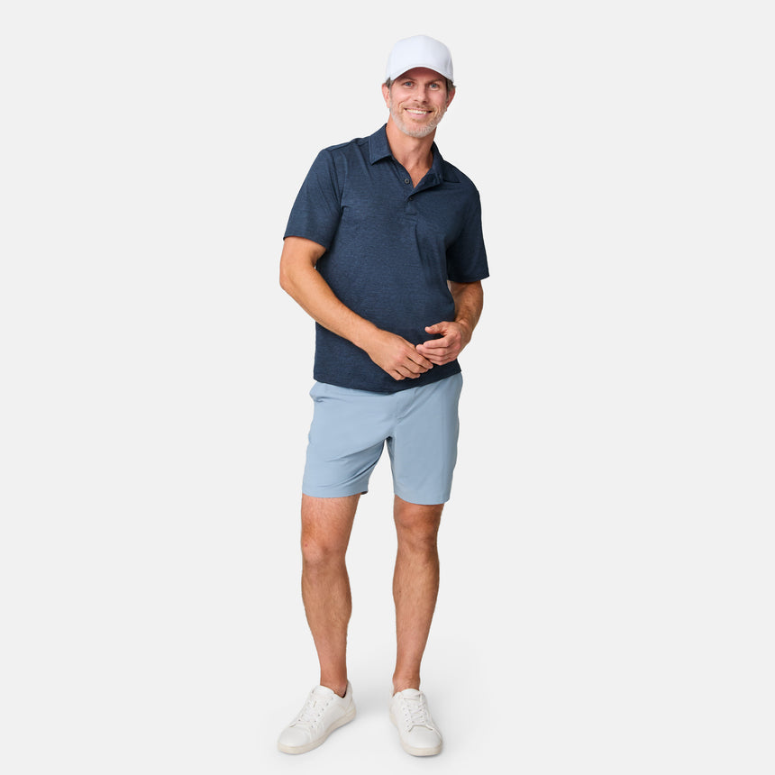 A man in a white cap, navy polo, Clearance Men’s Trek Short, and white sneakers stands smiling against a plain white background.