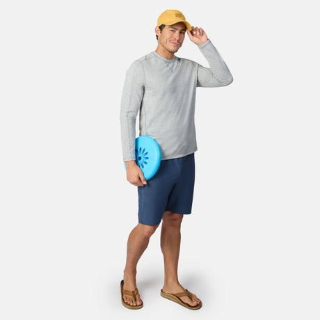 A man in a yellow cap, gray long-sleeve shirt, Clearance Men’s Trek Short, and sandals holds a blue frisbee and smiles against a plain white background.