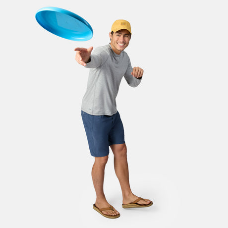 A smiling man in a yellow cap, gray long-sleeve shirt, and blue Clearance Men’s Trek Shorts throws a blue frisbee forward against a plain white background.