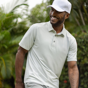 A man smiles outdoors among green plants, wearing a light gray Men's Tech Pique Polo Shirt from Clearance and a white cap.