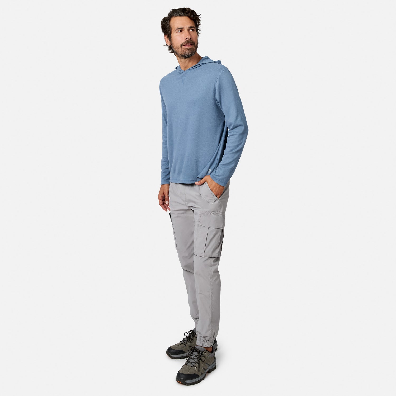 [Model is wearing size M.] A man wearing a dusty blue long-sleeve hooded shirt, light gray cargo pants, and hiking boots, standing against a solid off-white background.