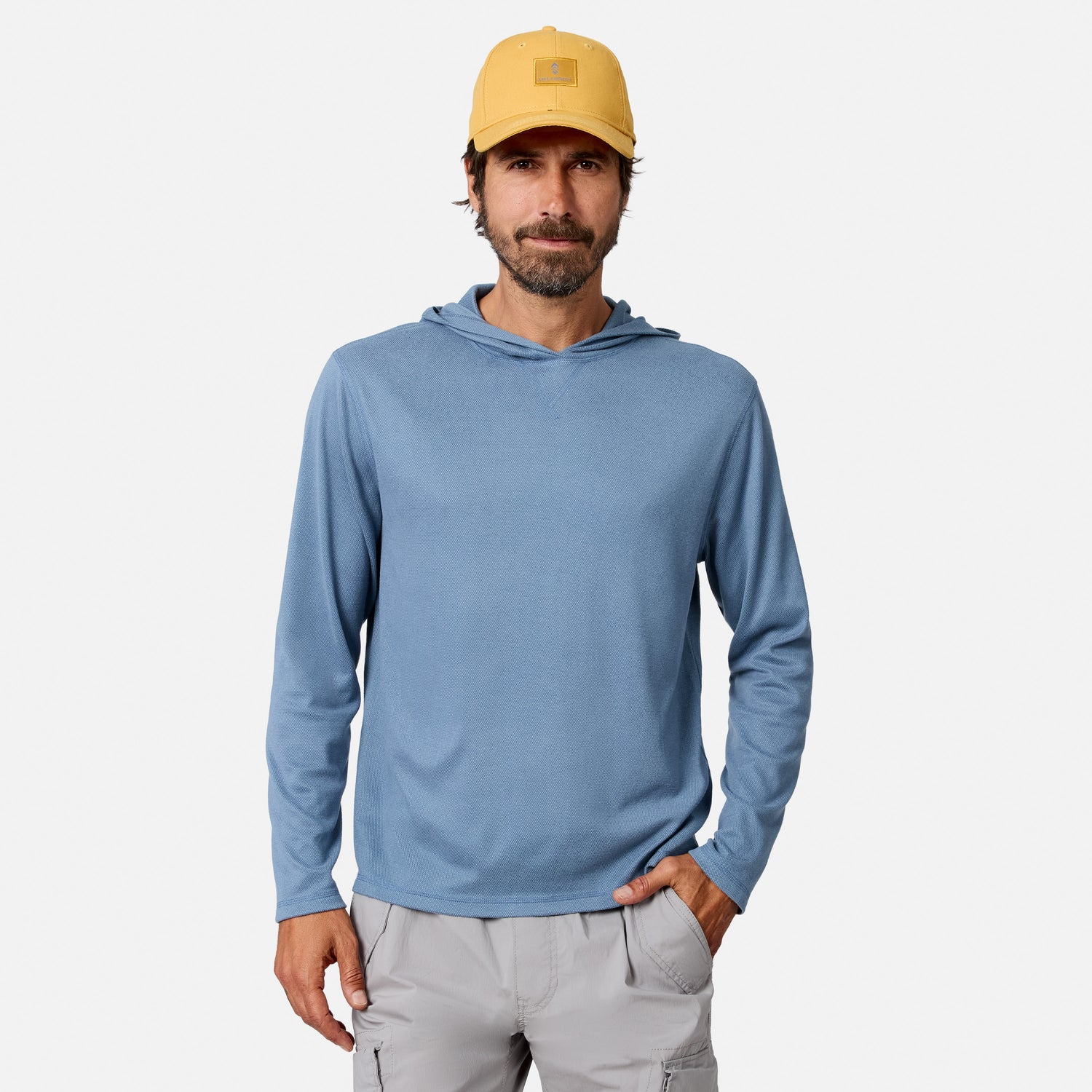 [Model is wearing size M.] A man wearing a light blue long-sleeve hooded shirt, a yellow baseball cap, and light gray cargo pants, standing against a plain light gray studio background.