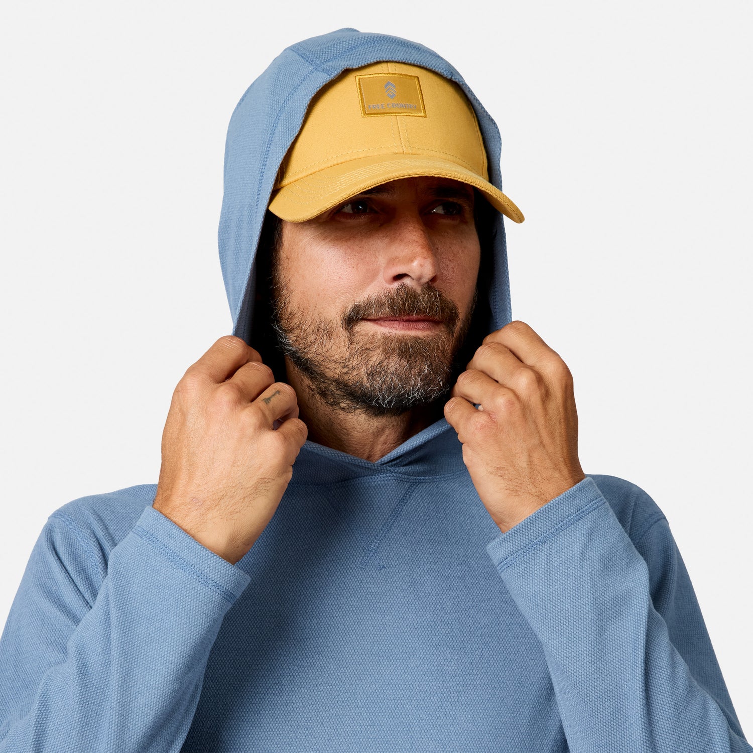 [Model is wearing size M.] A man wearing a light blue hoodie and a mustard yellow baseball cap, adjusting the hood of the sweatshirt against a plain light gray background.