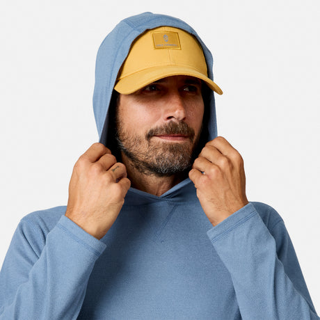 [Model is wearing size M.] A man wearing a light blue hoodie and a mustard yellow baseball cap, adjusting the hood of the sweatshirt against a plain light gray background.