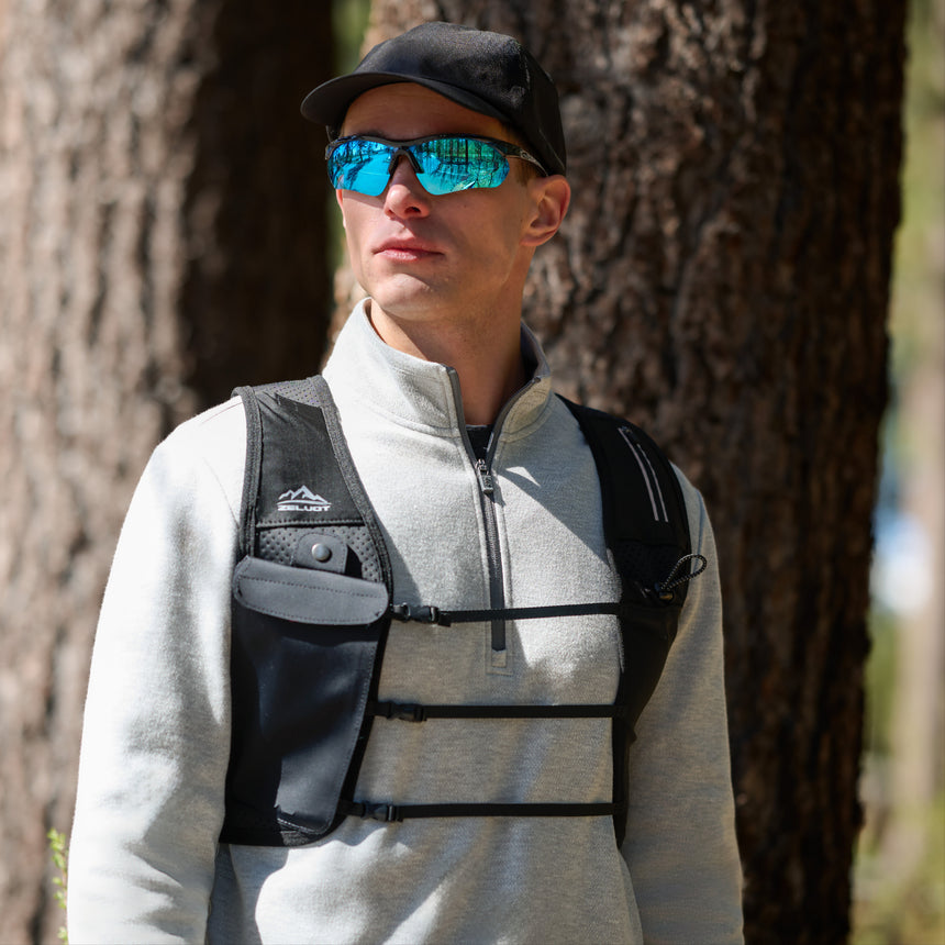 A man wearing a black cap, reflective blue sunglasses, and the Free Country Men’s Twill Interlock 1/4 Zip Long Sleeve Shirt in light gray with a black chest harness stands outdoors in a forest with tall trees.