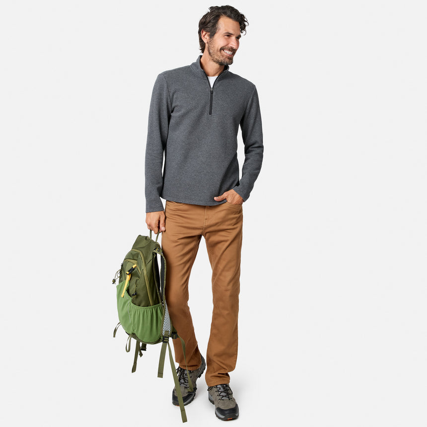 A man wearing the Free Country Men’s Twill Interlock 1/4 Zip Long Sleeve Shirt in gray, paired with tan pants and hiking shoes, holds a green backpack in his left hand while smiling and looking to his right.