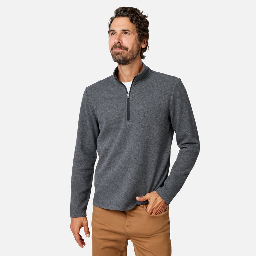 A man with dark hair and a beard wears the Free Country Men’s Twill Interlock 1/4 Zip Long Sleeve Shirt in Black Heather and tan pants, standing with one hand in his pocket against a plain white background.