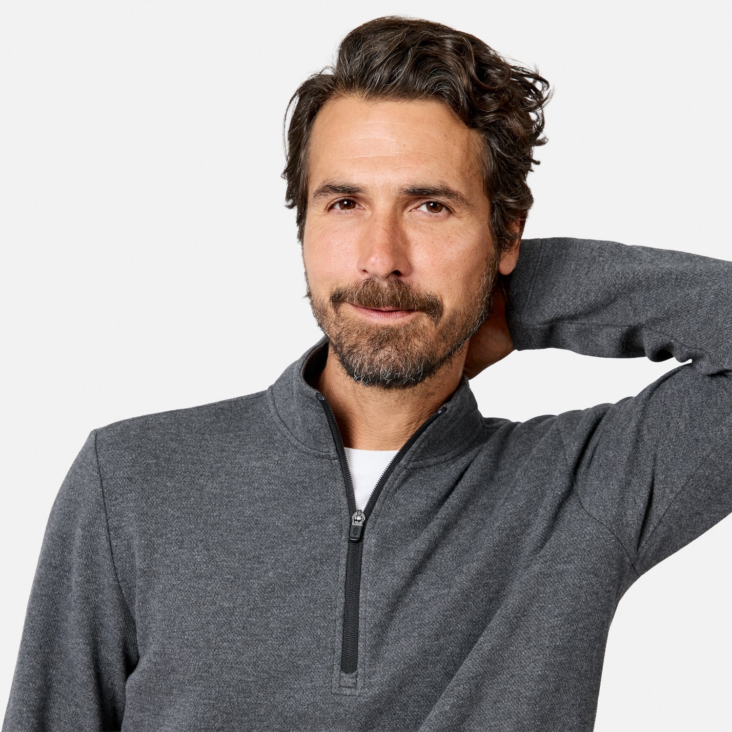 A man with dark hair and a beard is wearing the Free Country Men’s Twill Interlock 1/4 Zip Long Sleeve Shirt in gray over a white shirt. Made from breathable knit fabric, the quarter zip design adds both comfort and style. He stands against a plain light background, smiling slightly with one hand behind his head.