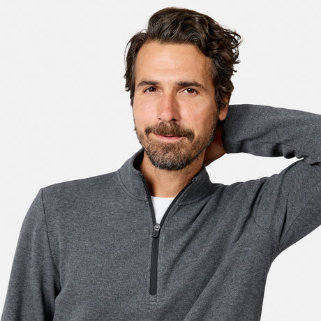 A man with dark hair and a beard is wearing the Free Country Men’s Twill Interlock 1/4 Zip Long Sleeve Shirt in gray over a white shirt. Made from breathable knit fabric, the quarter zip design adds both comfort and style. He stands against a plain light background, smiling slightly with one hand behind his head.