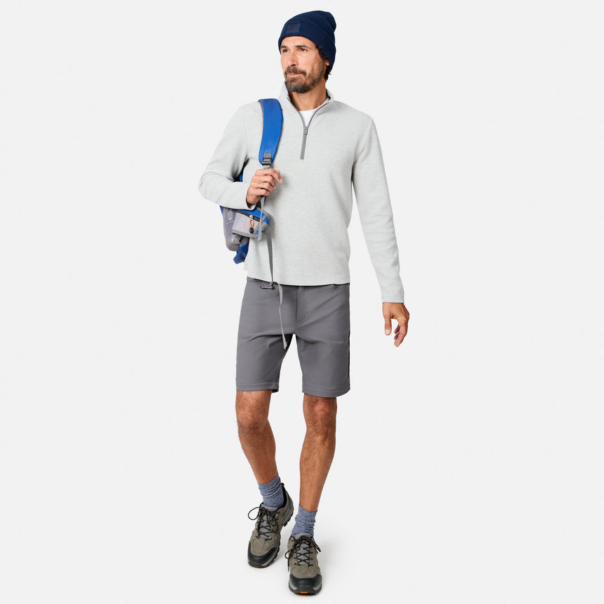 A man wearing a blue beanie, gray shorts, blue socks, hiking shoes, and carrying a blue-strap backpack on his shoulder models the Free Country Men’s Twill Interlock 1/4 Zip Long Sleeve Shirt against a plain white background.