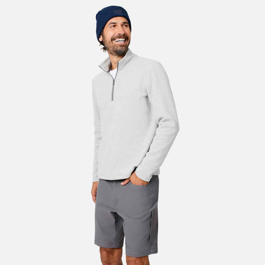A smiling man stands with one hand in his pocket against a plain white background, wearing a navy beanie, grey shorts, and the Free Country Men’s Twill Interlock 1/4 Zip Long Sleeve Shirt in light grey, crafted from breathable knit.