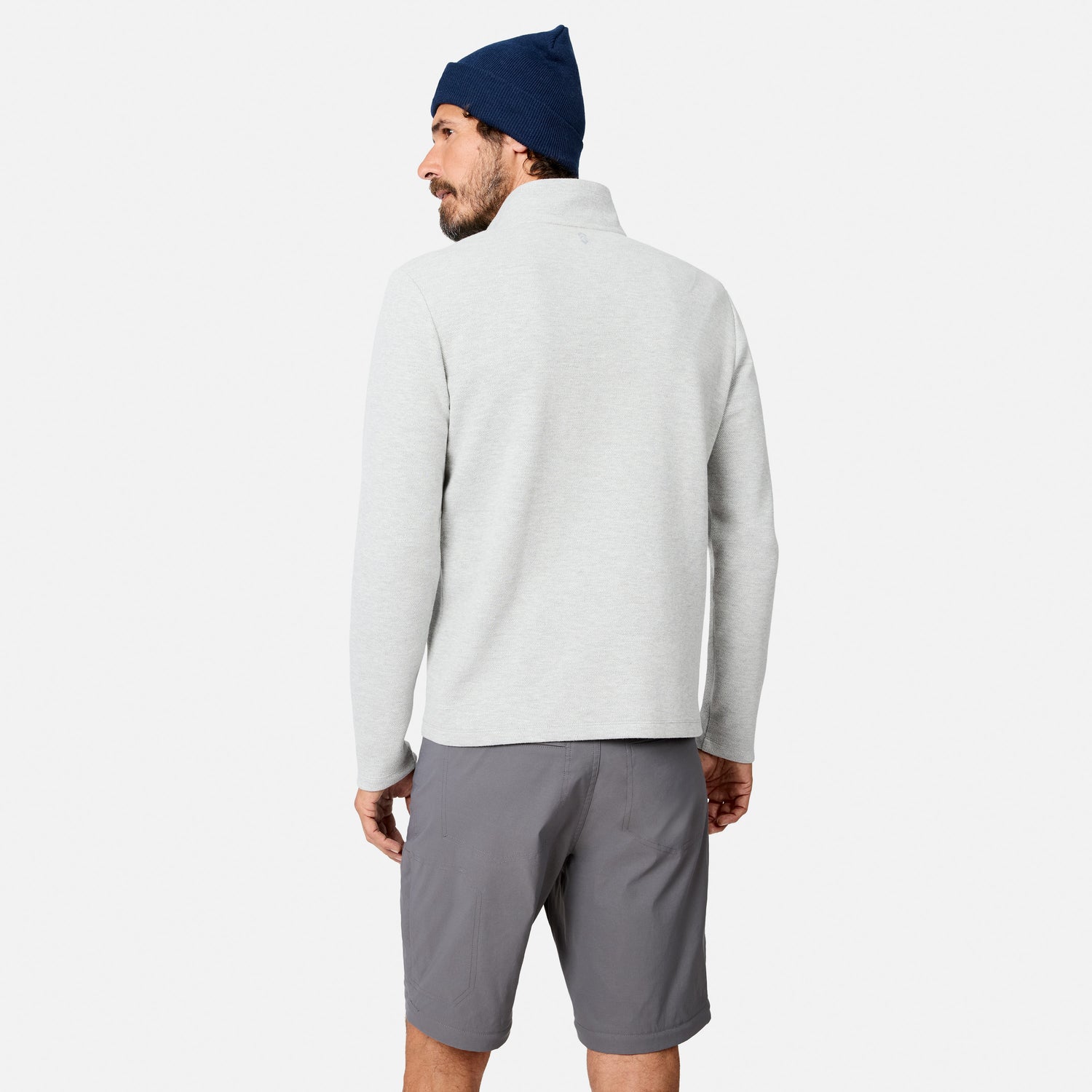 A man stands facing away against a plain white background, wearing a light gray Men’s Twill Interlock 1/4 Zip Long Sleeve Shirt by Free Country, paired with dark gray shorts and a navy beanie.